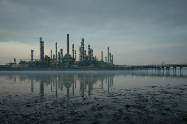 An estuary chemical works at dusk with tall distillation columns and a reformer stack standing dark and unlit, a single flare extinguished, mist over the tidal mudflats of a wide river