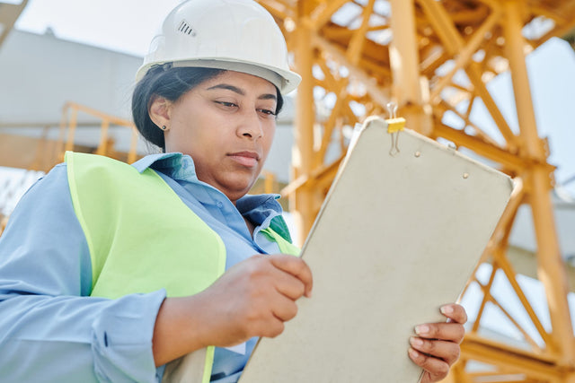 Confident female construction worker inspecting site with clipboard and helmet.