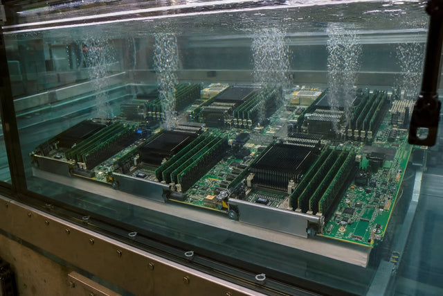 A server circuit board fully submerged in clear dielectric fluid with fine bubble streams rising from warm components.