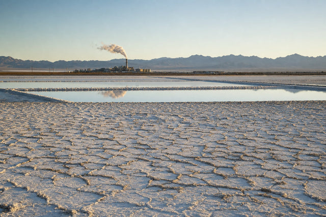 A dry alkali lakebed at golden hour: cracked white salt crust in the foreground, shallow brine evaporation ponds behind it, and the silhouette of a chemical works with a steaming stack against desert mountains