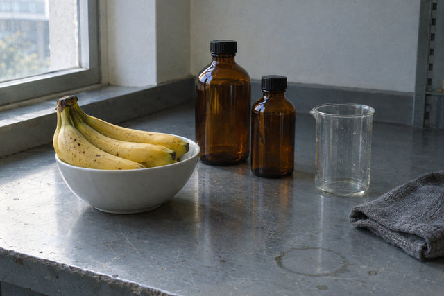Ripe bananas in a white bowl beside two unlabelled amber laboratory bottles and a glass beaker on a scratched steel bench, lit by window daylight — the fruit and the industrial solvent that share the ester isoamyl acetate.