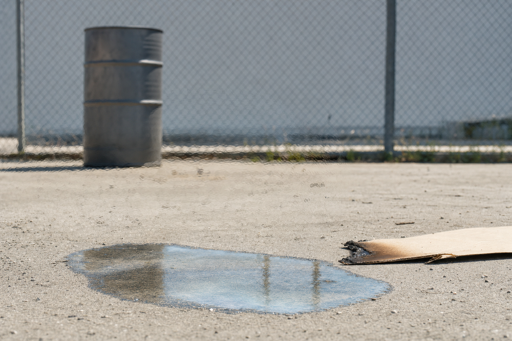 Heat shimmer bending a chain-link fence above a clear puddle in a steel tray, with scorched cardboard beside it and no visible flame