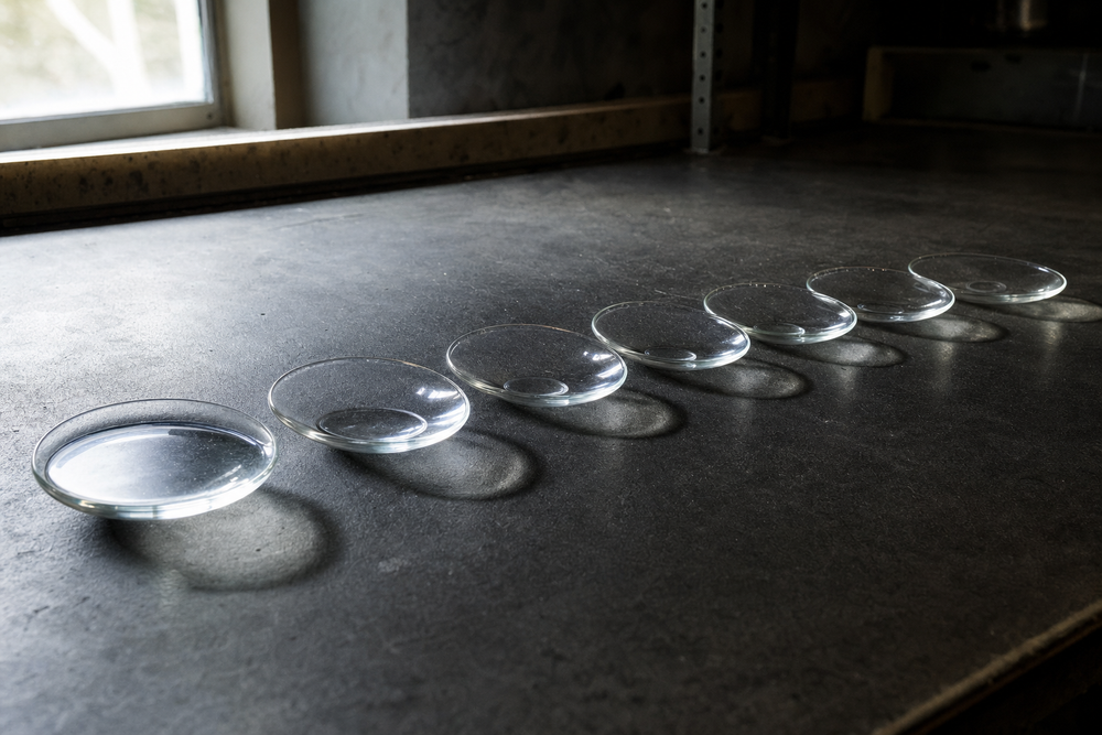 Seven identical unlabeled glass dishes in a row on a dark laboratory bench, each holding progressively less clear solvent, illustrating a relative evaporation rate scale.