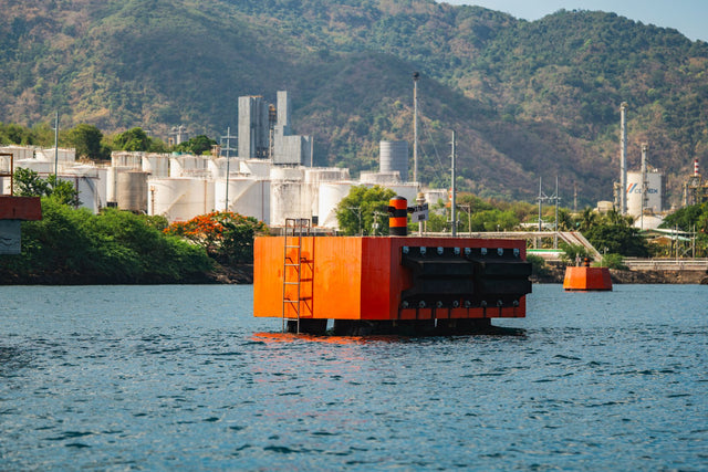 Orange floating buoy in a water body near an industrial facility with mountains in the background.