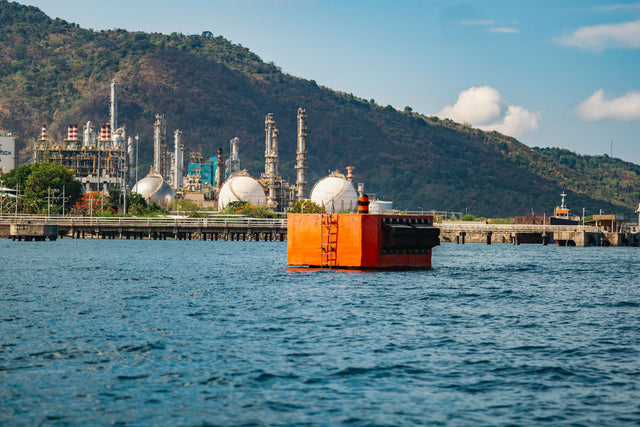 Industrial complex with orange buoy on water, near lush hills under a blue sky.