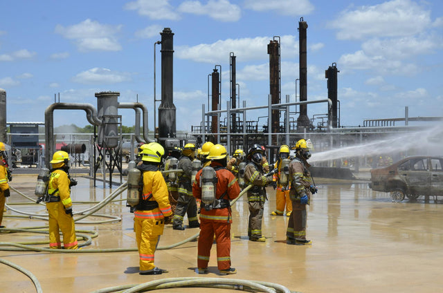 Firefighters wearing protective gear conduct a safety drill at an industrial site with heavy equipment.