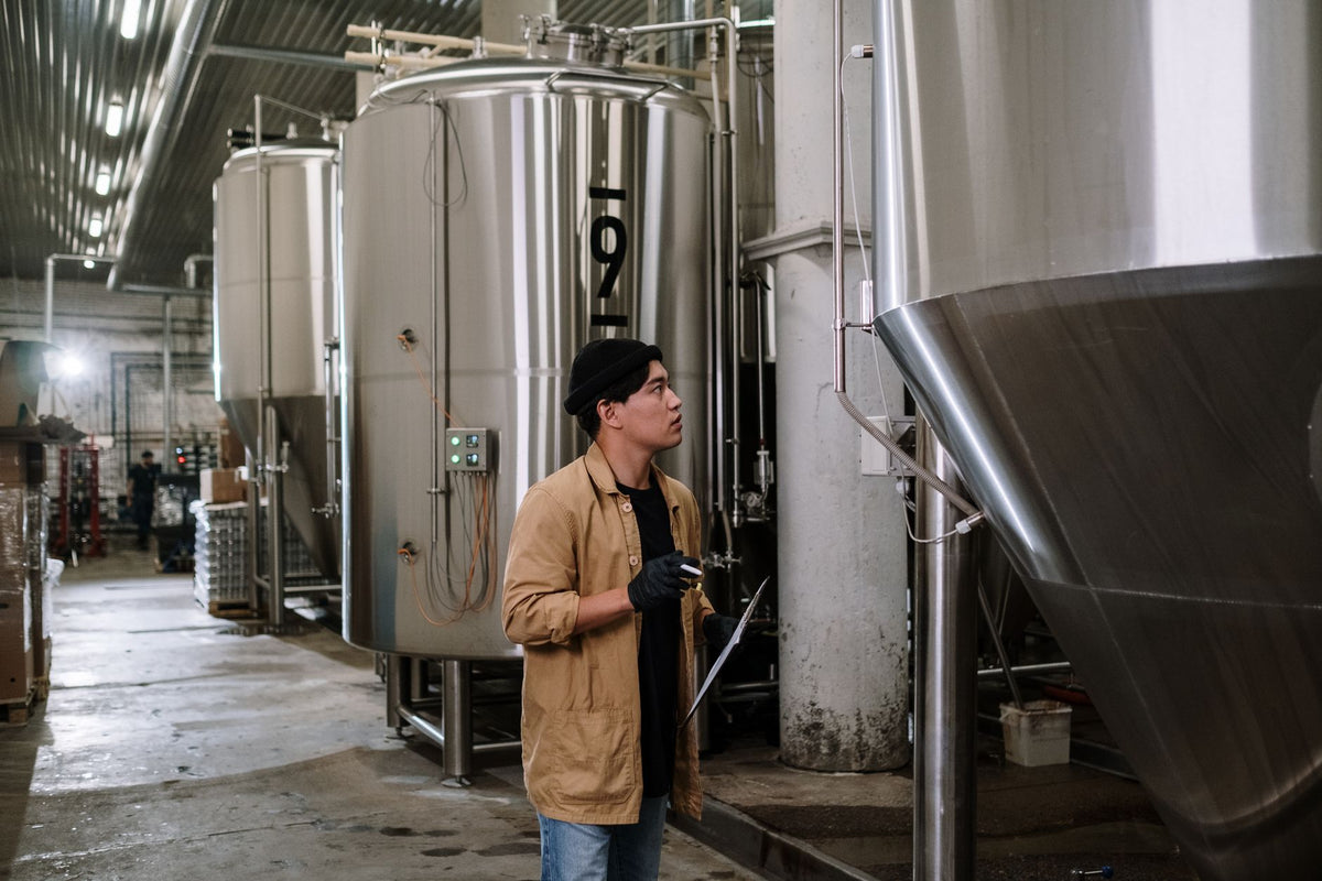 Asian man inspecting stainless steel tanks in a brewery, showcasing modern industrial processes.