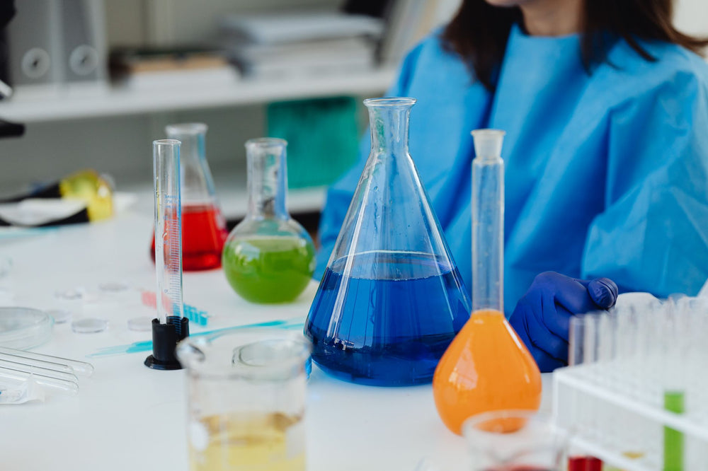 A close-up of a laboratory desk with colorful chemicals in glassware, featuring a researcher in protective gear.