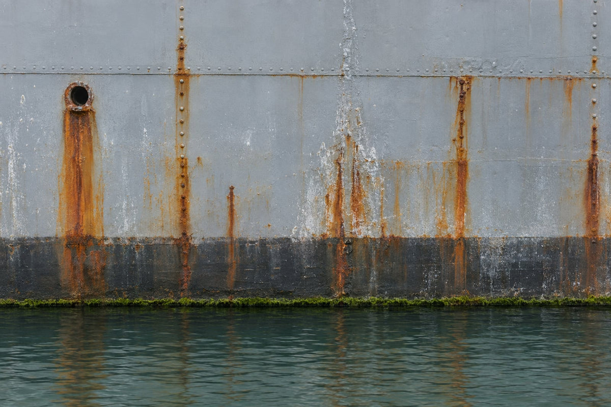 Weathered grey steel ship plating at the waterline: dark, nearly untouched steel below the line and long streaks of orange running rust in the splash zone above it