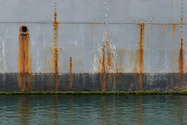 Weathered grey steel ship plating at the waterline: dark, nearly untouched steel below the line and long streaks of orange running rust in the splash zone above it