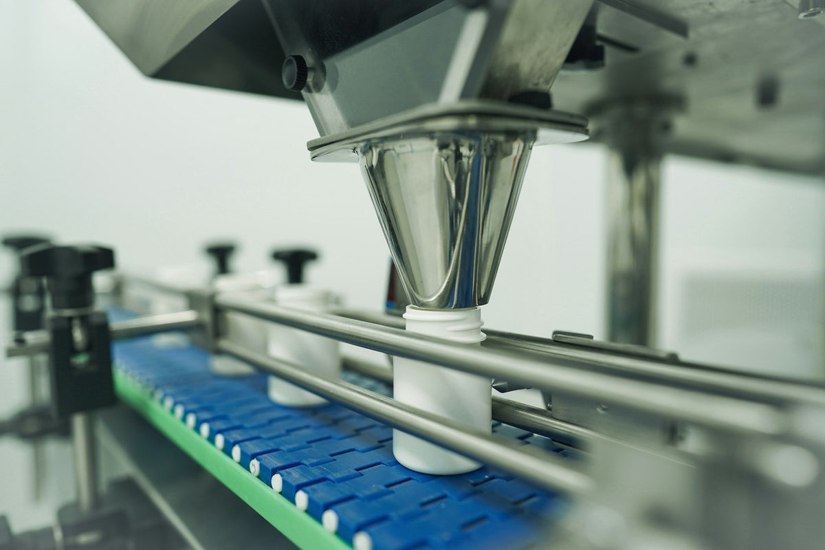 White plastic bottles on a blue conveyor passing under a stainless-steel filling funnel
